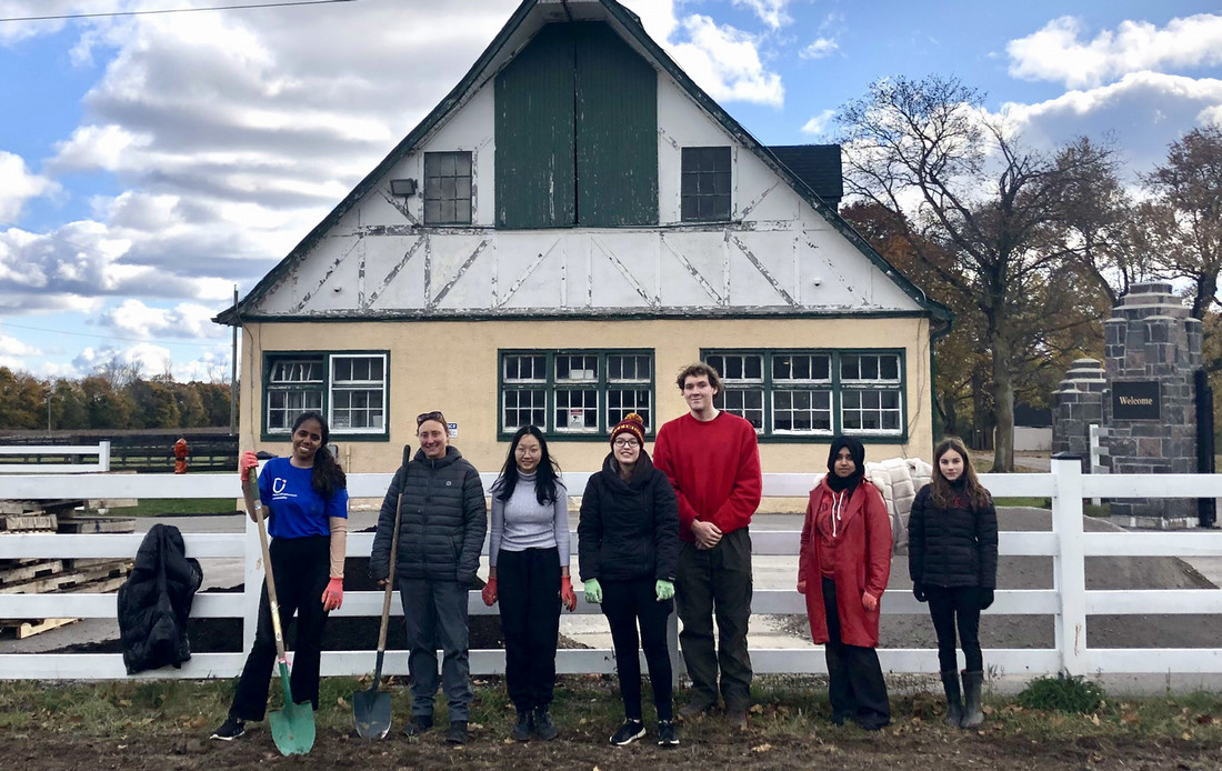 A group of people stands together in front a white fence and building on Windfields Farm.