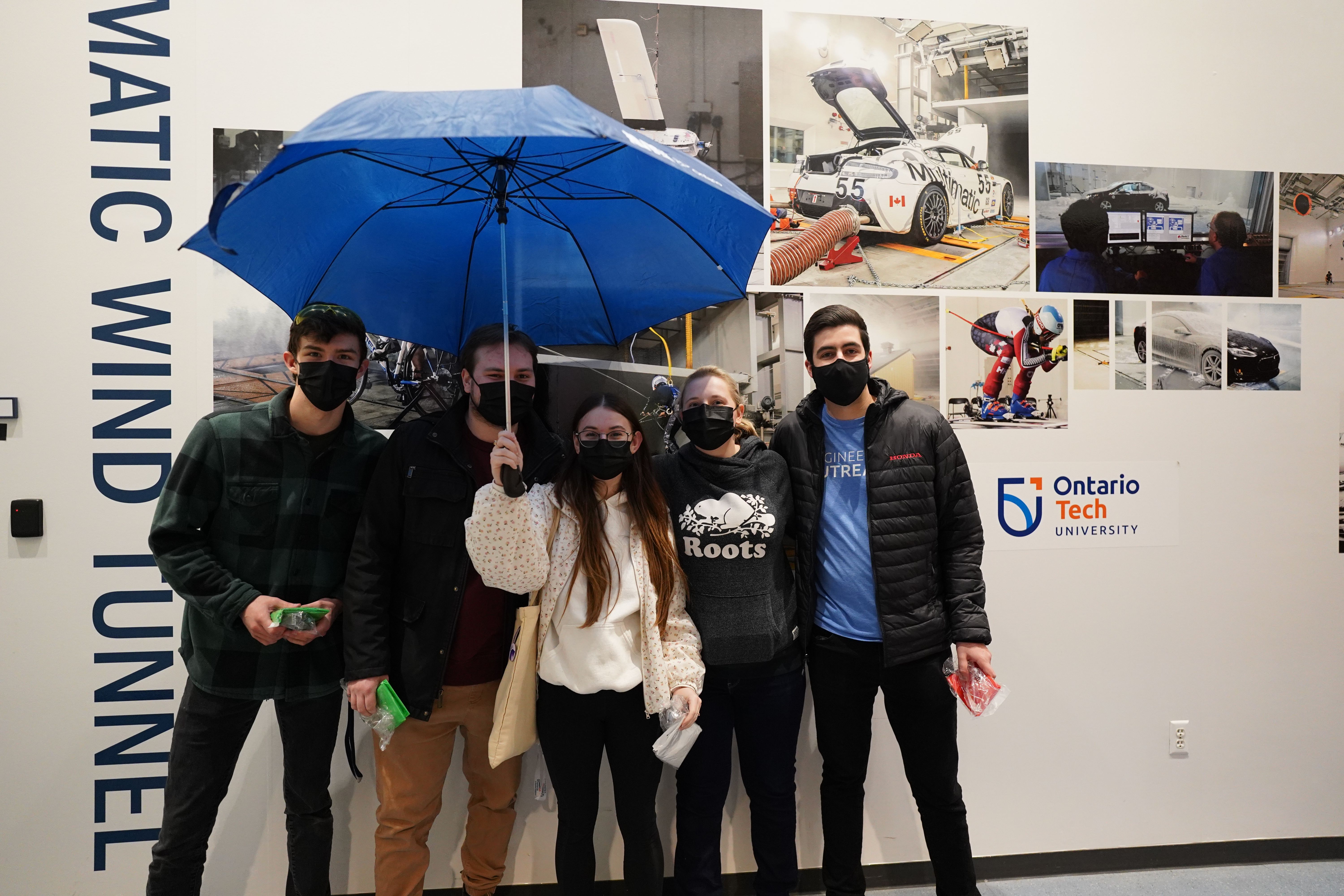 Five students are standing in front of a photo wall outside the climatic wind tunnel with a blue umbrella.  ${altNumber}