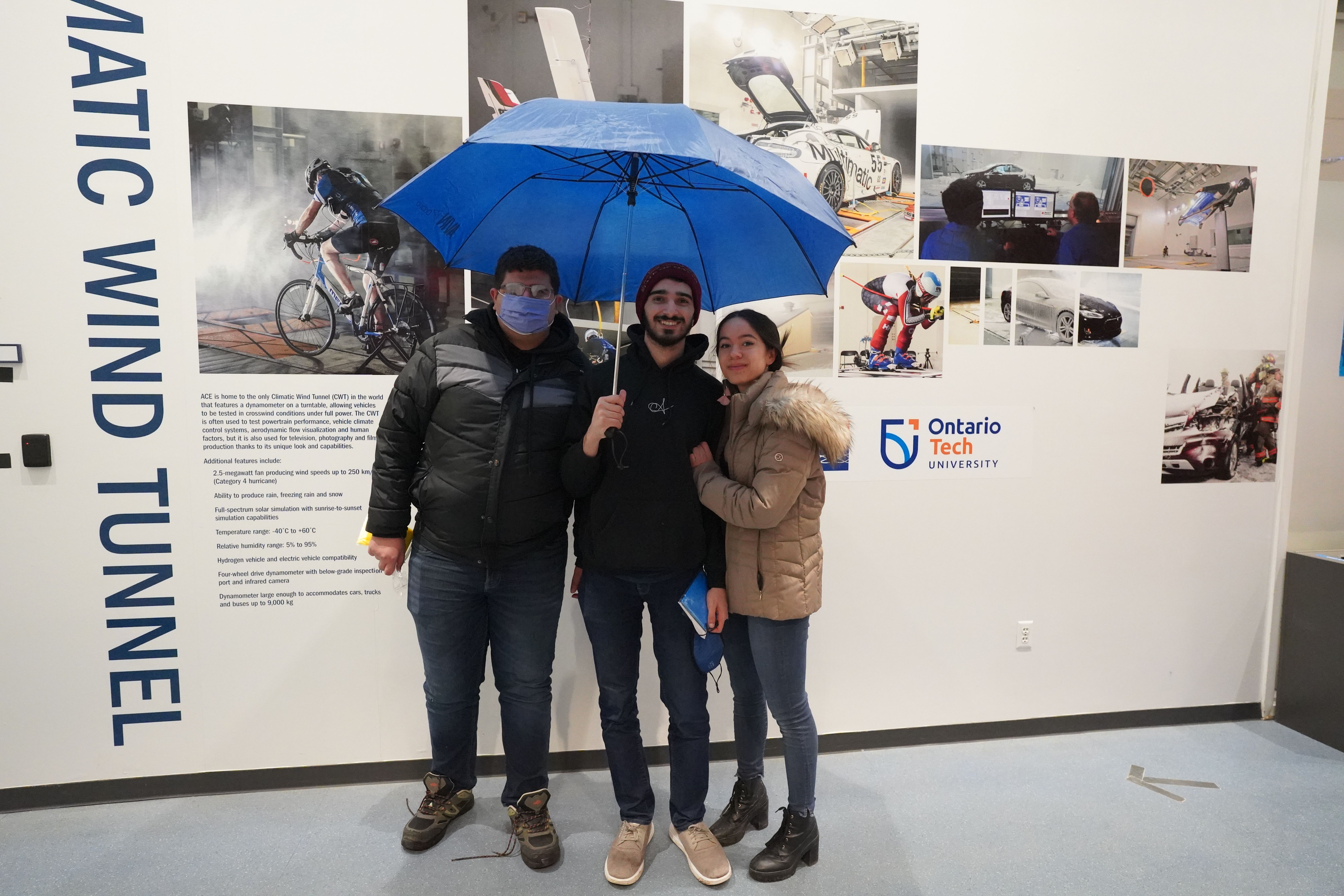 Three students are standing in front of a photo wall outside the climatic wind tunnel with a blue umbrella.  ${altNumber}