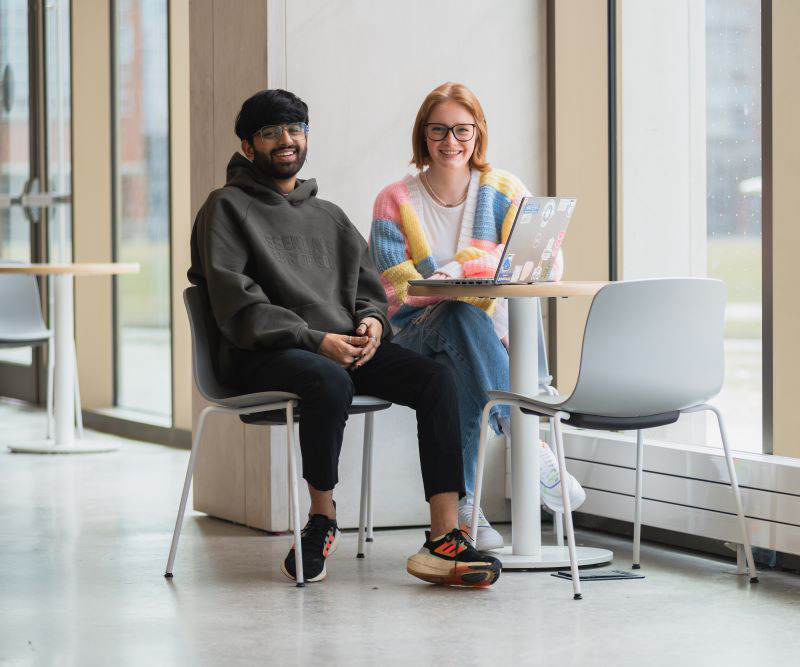 Two students sit at a table in Shawenjigewining Hall, smiling.