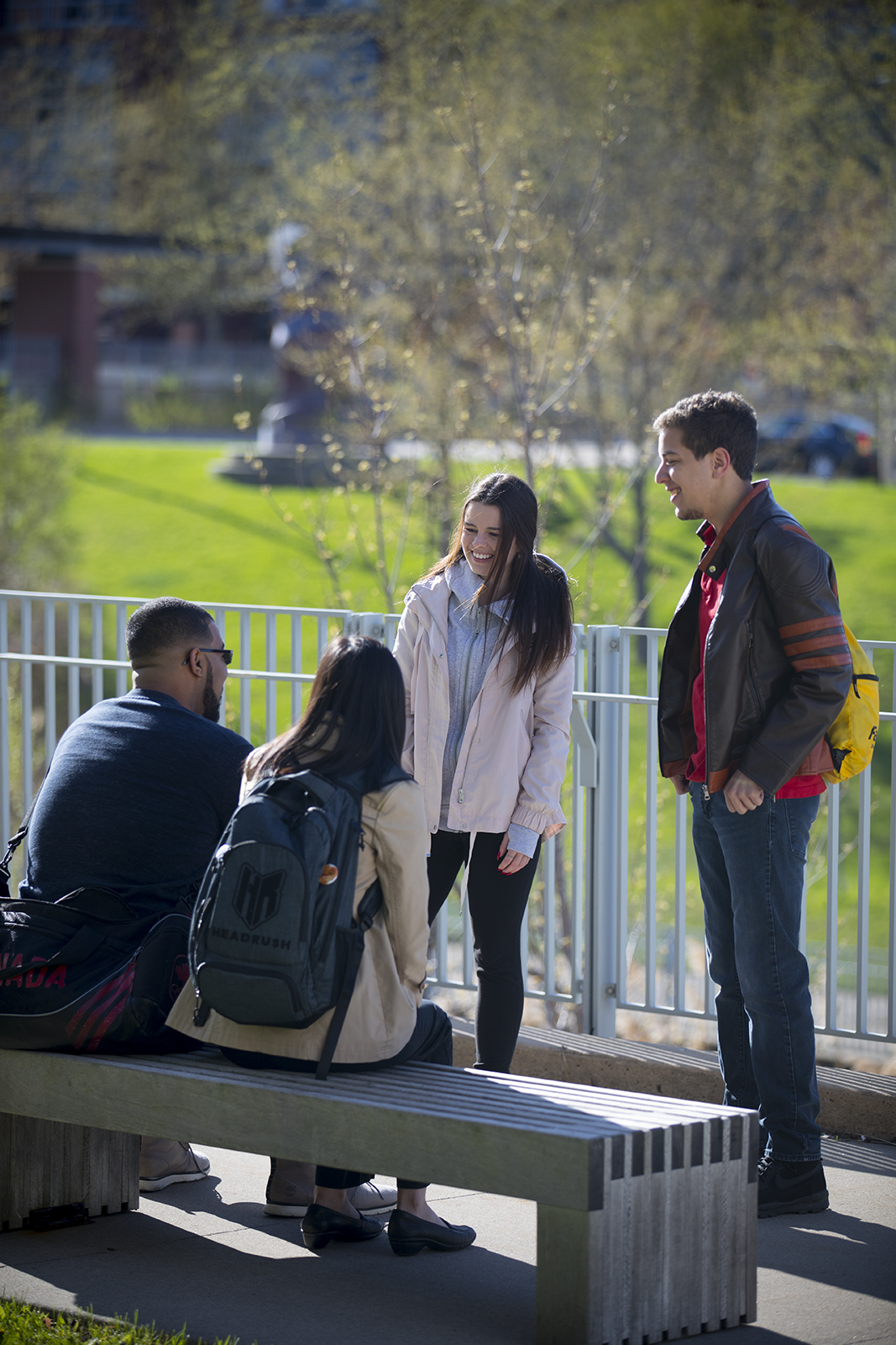 Four students sitting and standing outside on campus and smiling at each other.