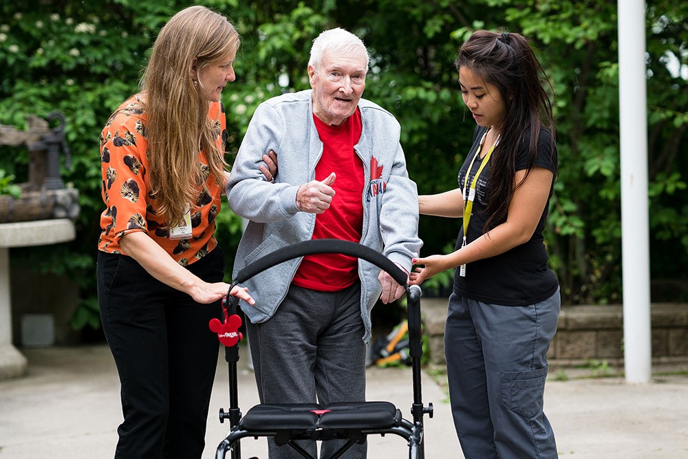 Two care givers stand on each side of a senior with a walker.