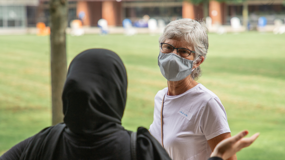 A student wearing a dark hijab with her back to the camera talks to an older adult, who is wearing a mask. 