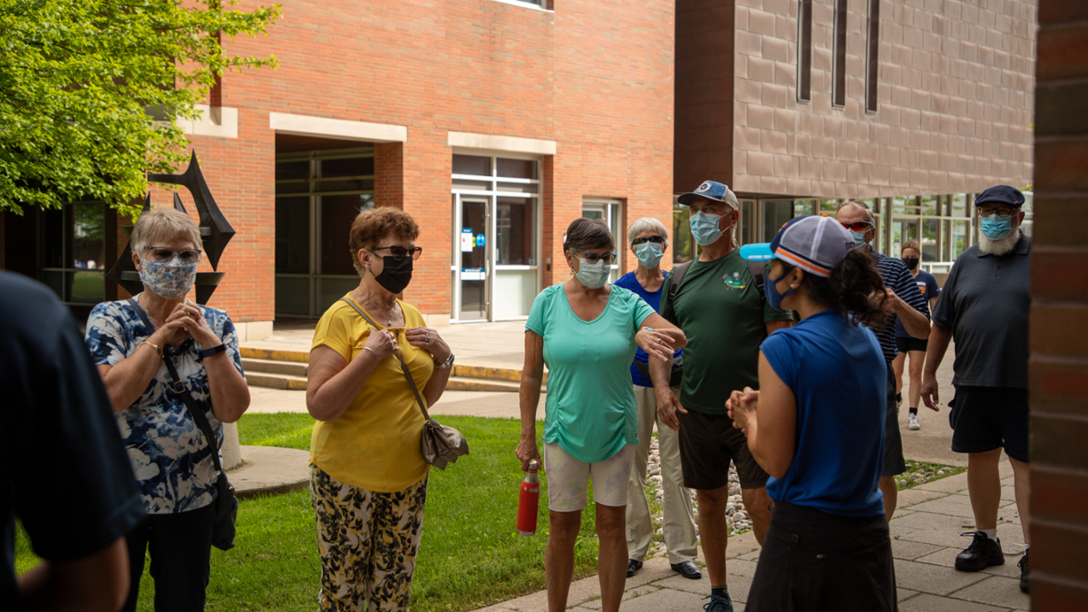 A group of older adults stand outside of orange brick buildings wearing masks