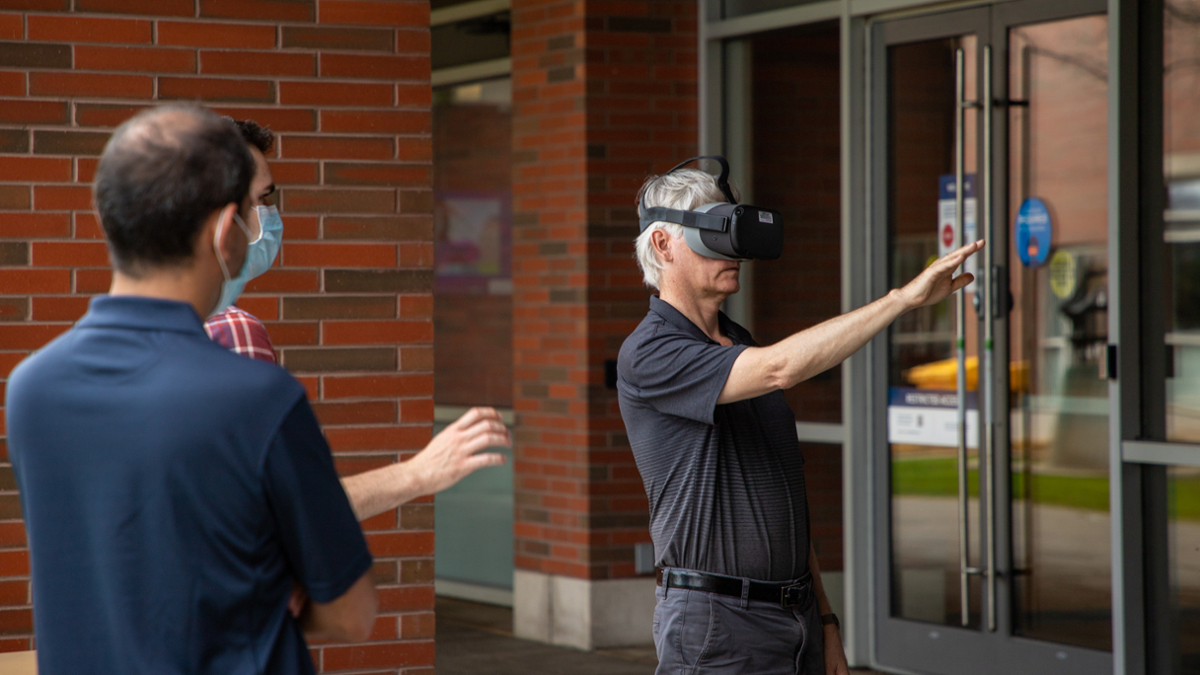 an older adult wears virtual reality goggles as others look on and instruct him in their use. 