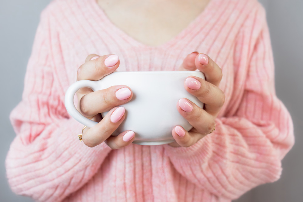 woman in a pink sweater and pink nails holds a white cup