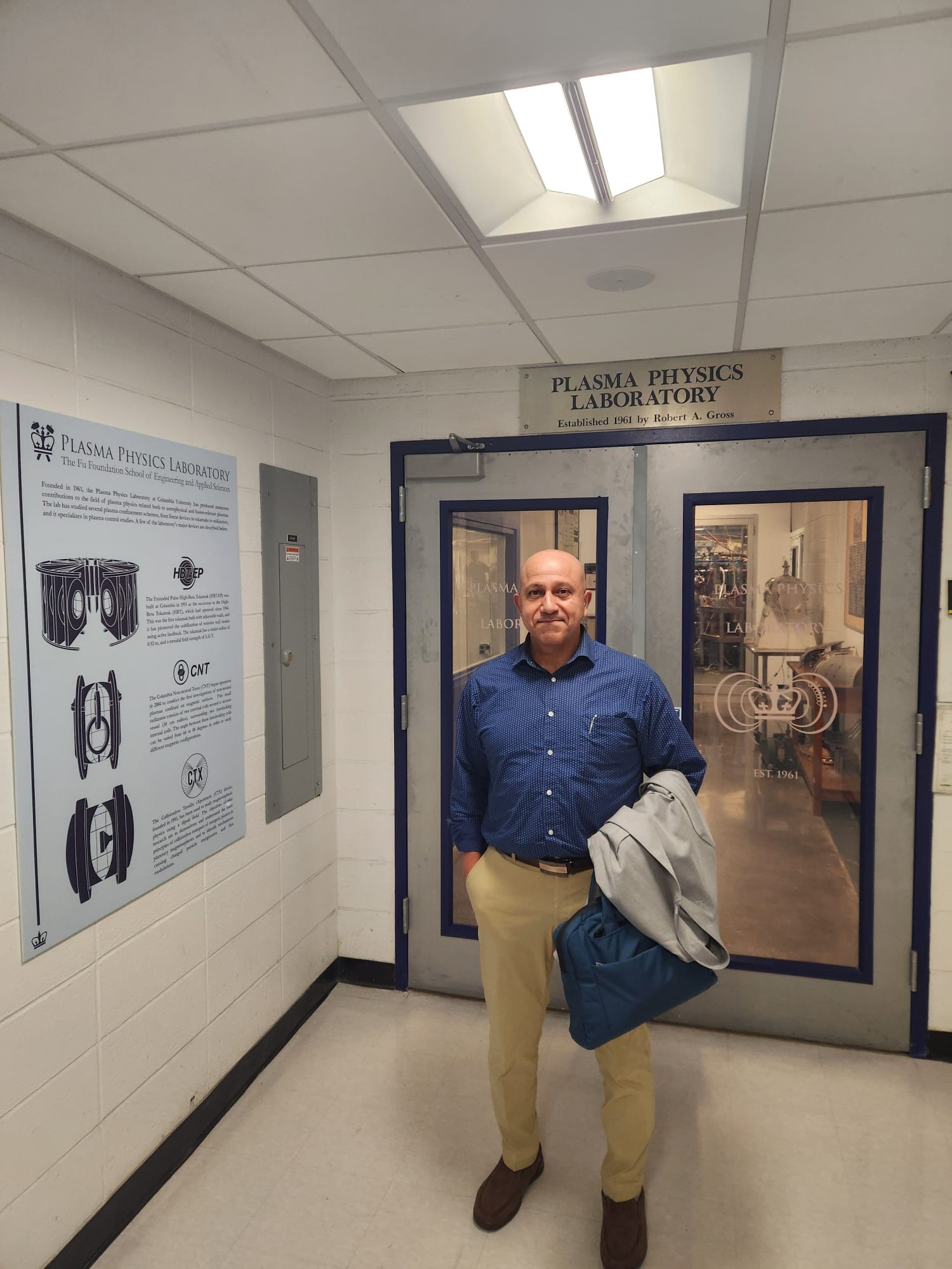 A man stands in front of the Plasma Physics Laboratory entrance, wearing a blue shirt and beige pants, holding a jacket.