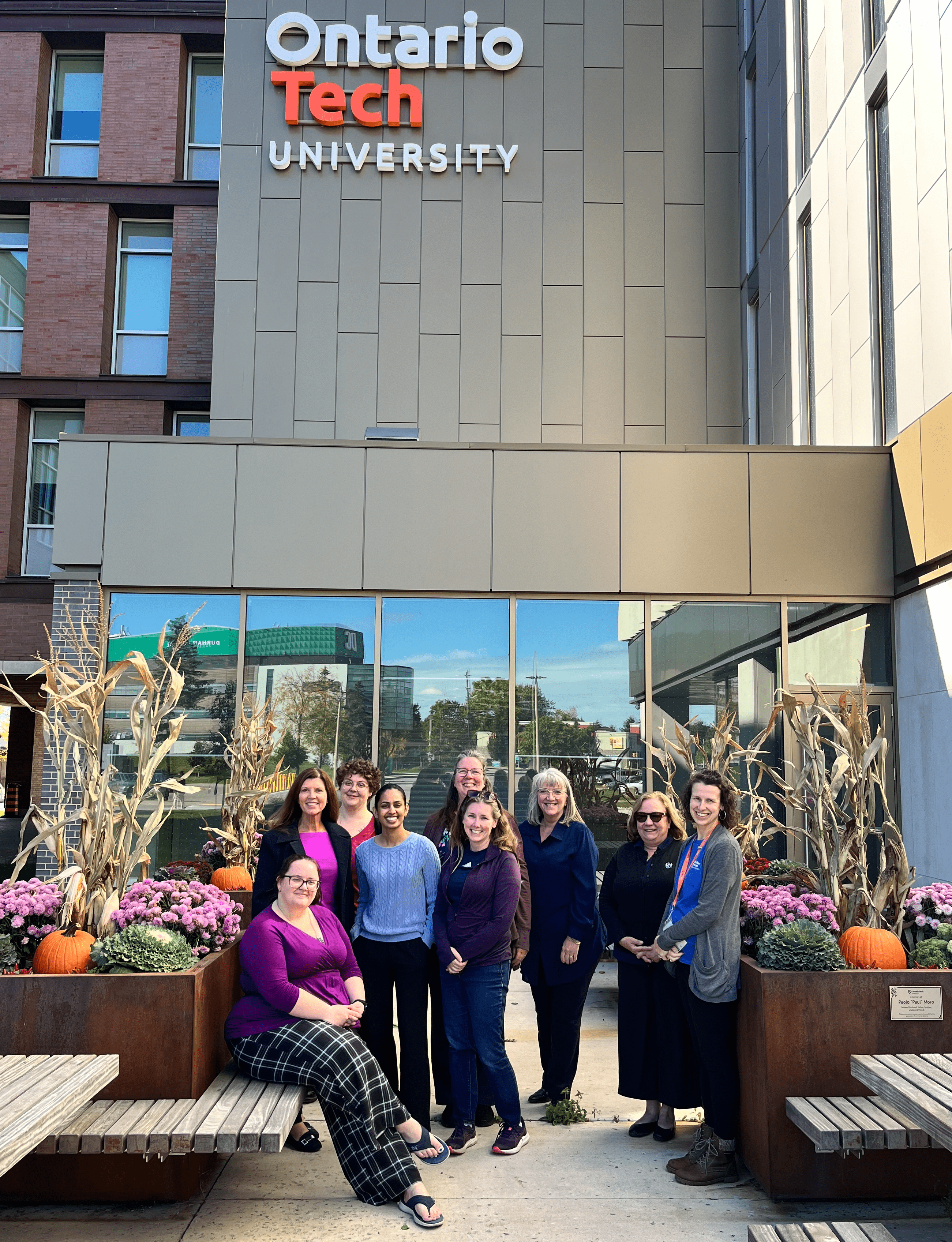 Ontario Tech University staff posing outside the campus building, dressed in blue and purple, surrounded by pumpkins and fall flowers.