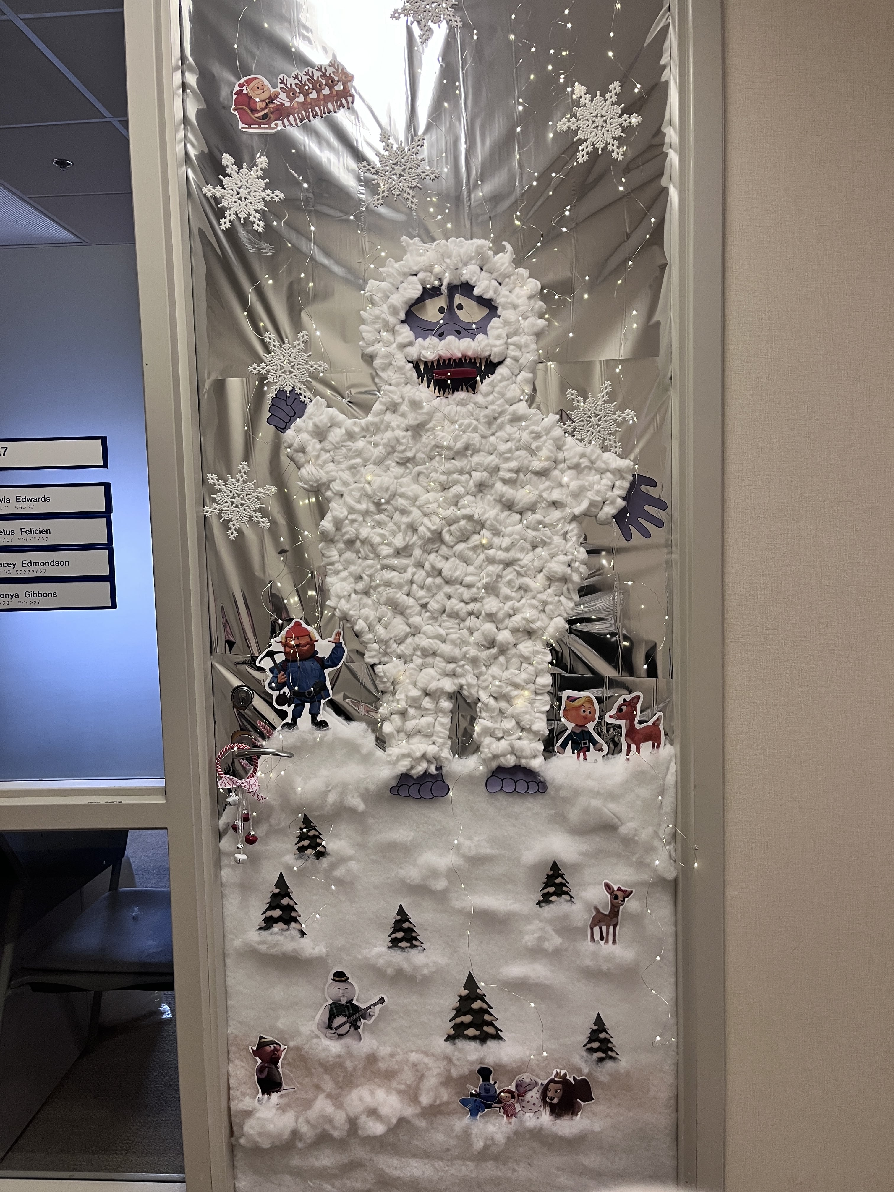 A whimsical winter-themed decoration featuring a fluffy yeti made from cotton balls, surrounded by snowflakes, trees, and cheerful cartoon characters.