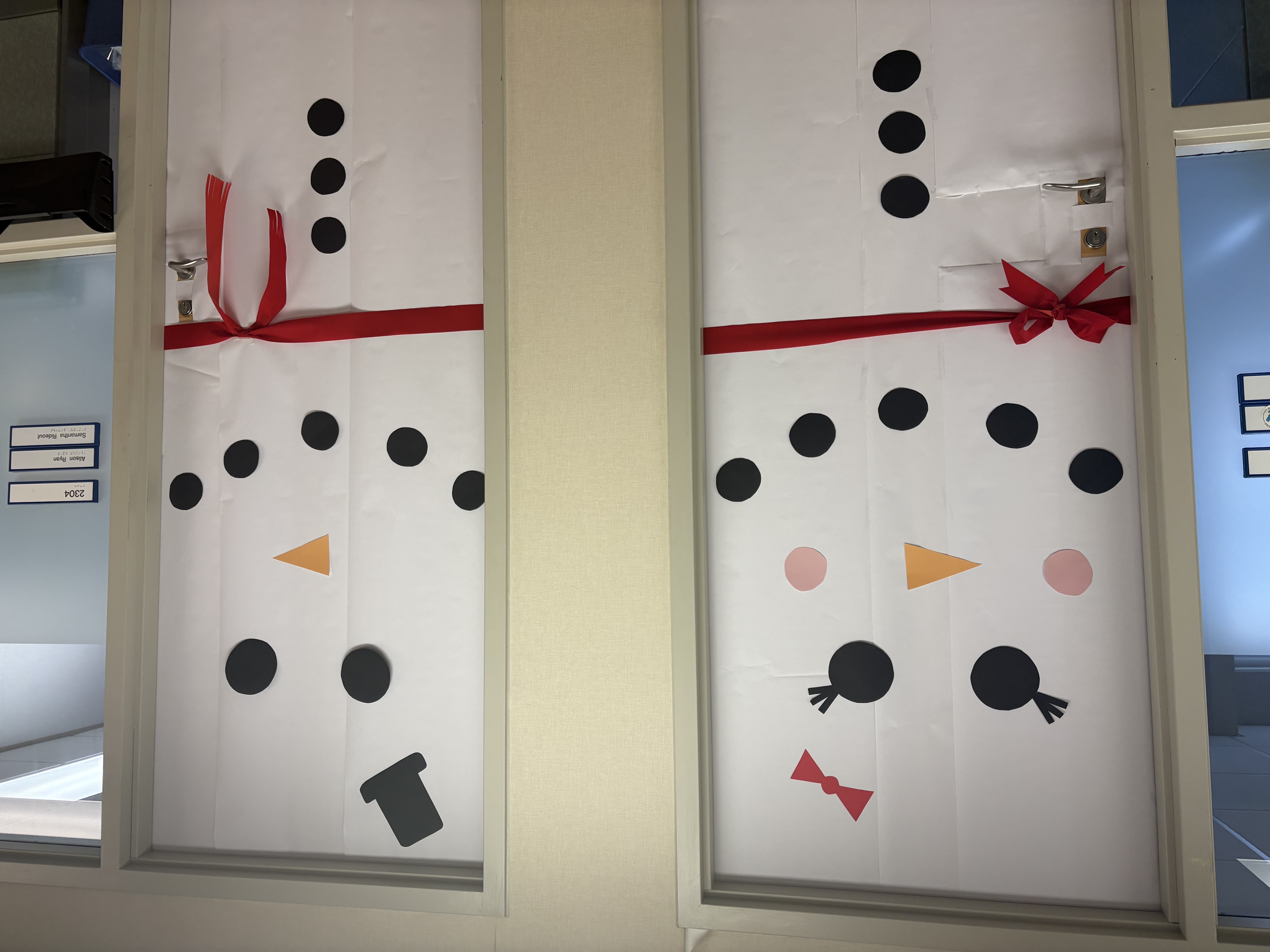 Decorative snowman-themed doors adorned with paper cutouts, featuring a black top hat, a red bow, and orange carrot noses.