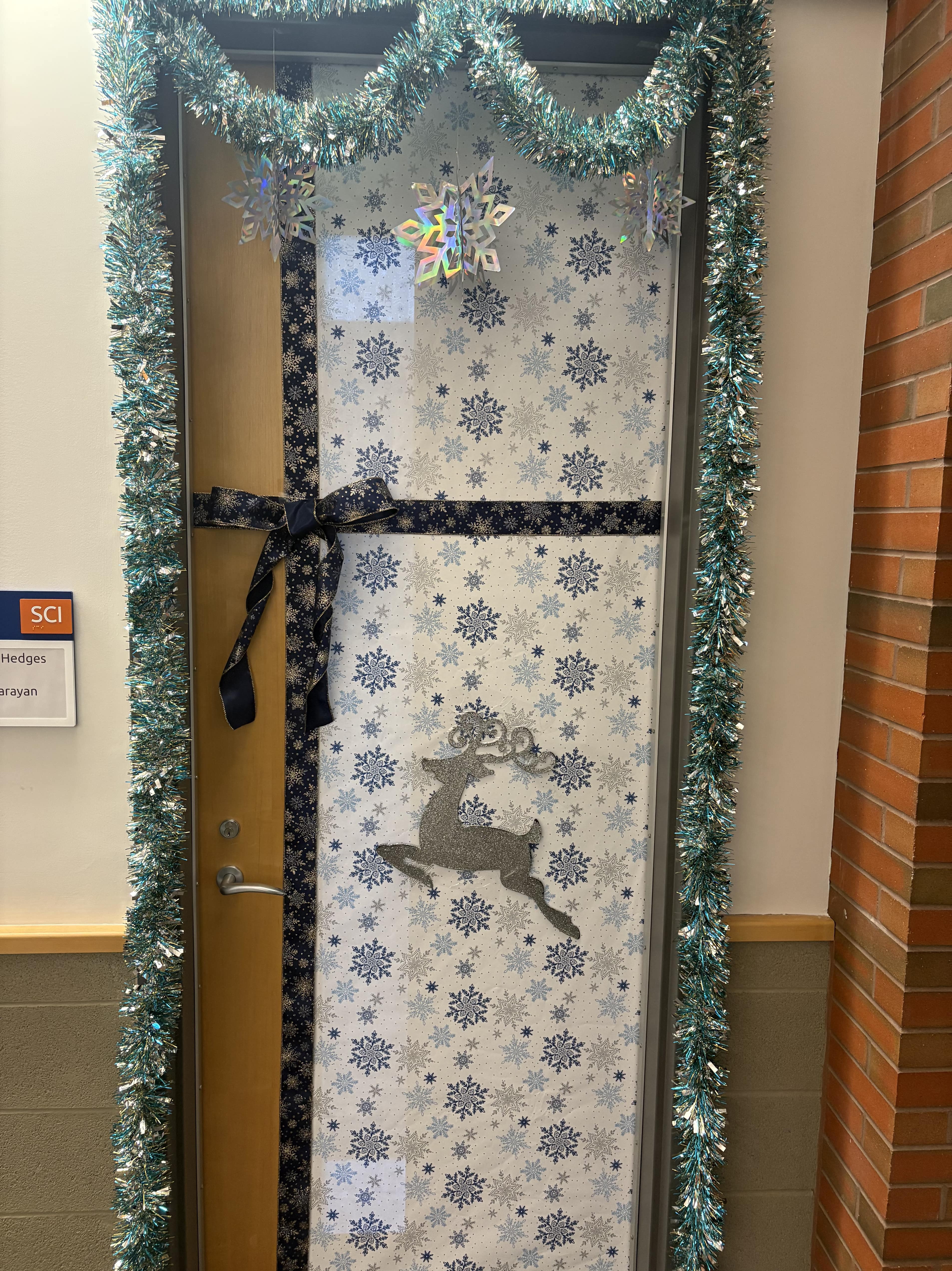 A festive holiday-decorated door featuring blue and white snowflake wrapping paper, adorned with a reindeer cutout and tinsel garland.