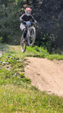 Samantha Woitzik riding a mountain bike over a jump on a forest trail while wearing a helmet and protective gear.