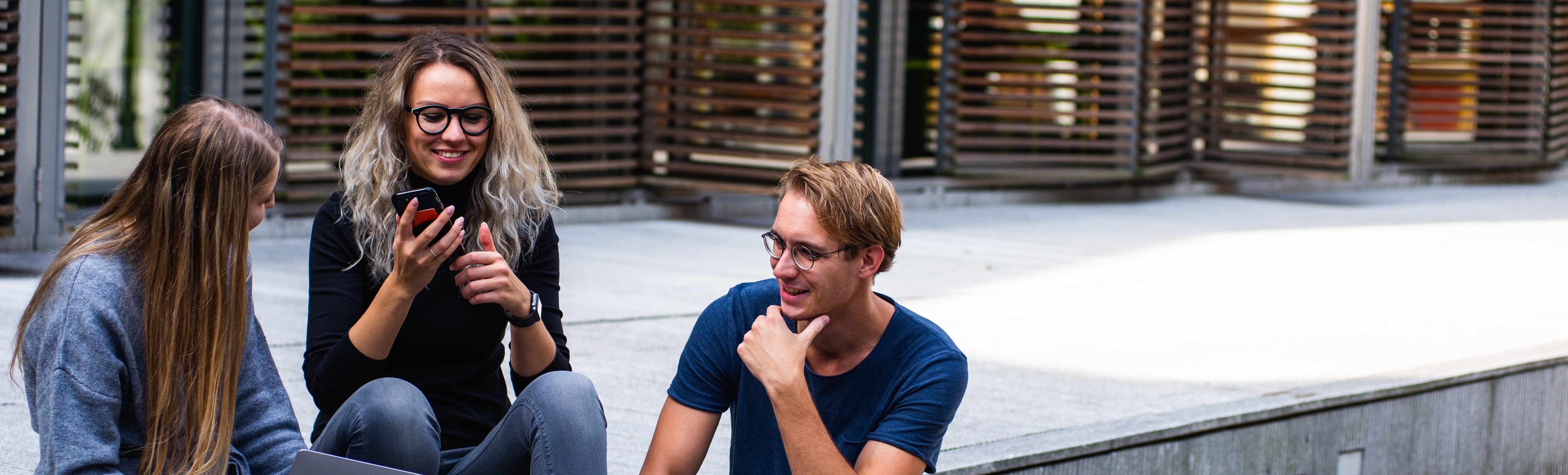 Three students seated outdoors on steps, looking at mobile devices. Photo by Buro Millennial from Pexels.