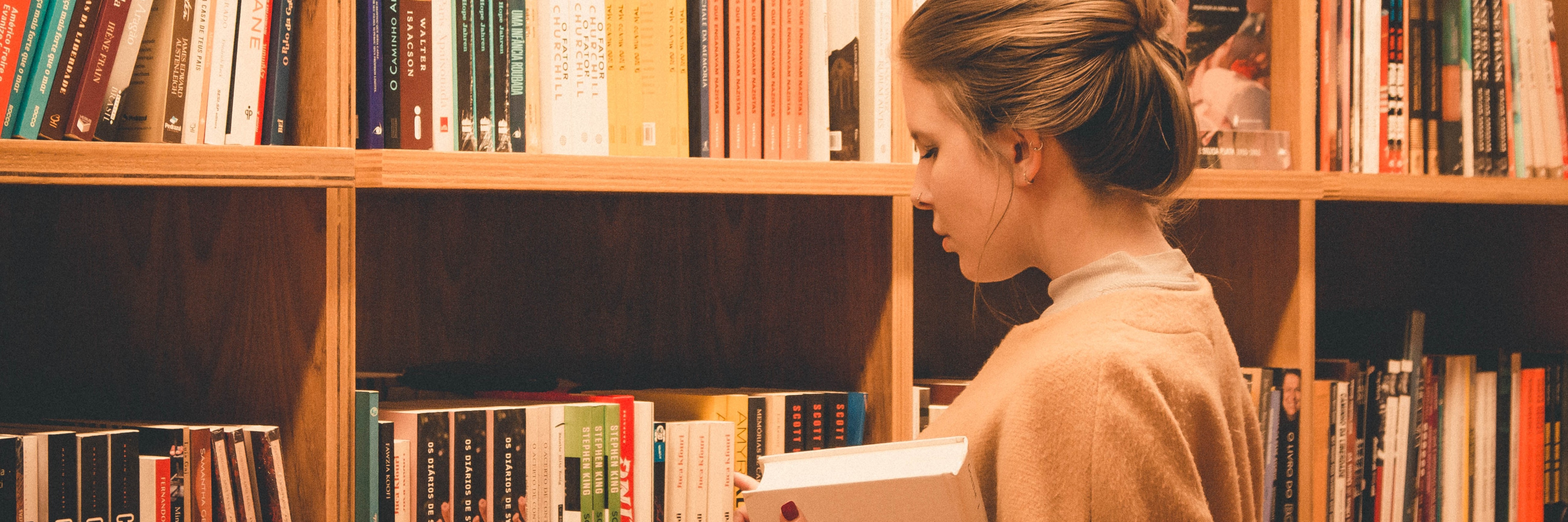 A woman holding a book, looking at a library shelf full of books. Photo by Eliabe Costa on Unsplash