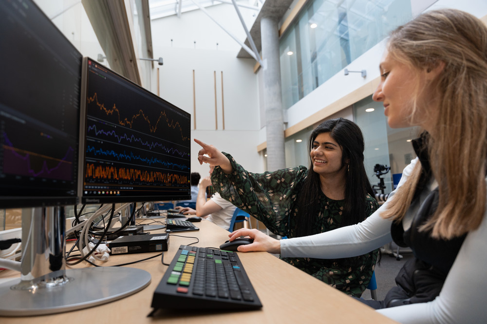 Two students collaborate in a modern computer lab, analyzing financial graphs on dual monitors