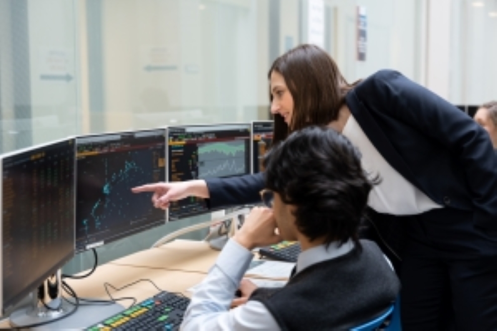 two people looking at multiple computer monitors. One person is pointing at a graph on the middle monitor