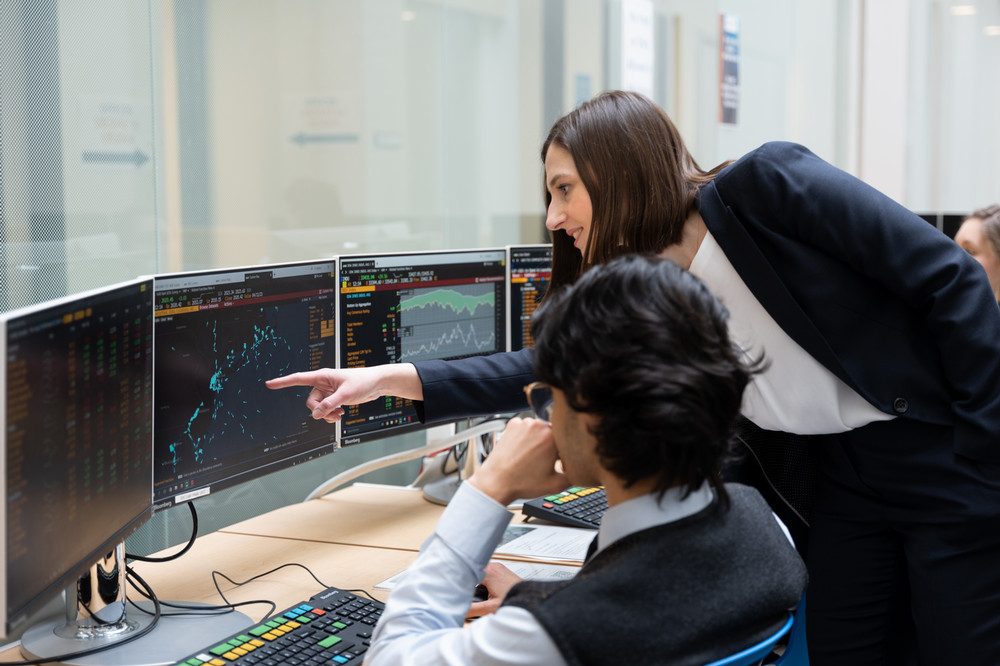 two people looking at multiple computer monitors. One person is pointing at a graph on the middle monitor
