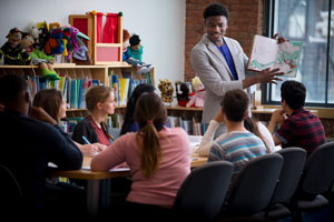 students at table with adult standing and holding a book