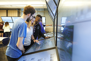 A group of students stand closely over a series of monitors engaging with data