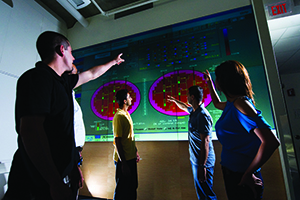 A male instructor points something out on a large diagram on the wall to a group of students