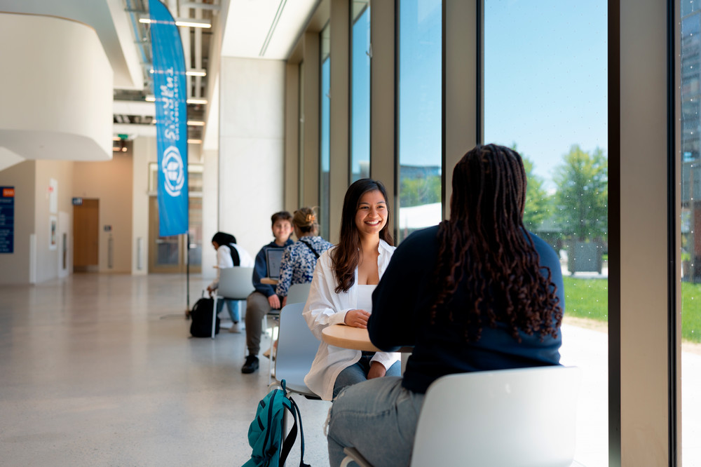 Students sitting in common area by windows. 