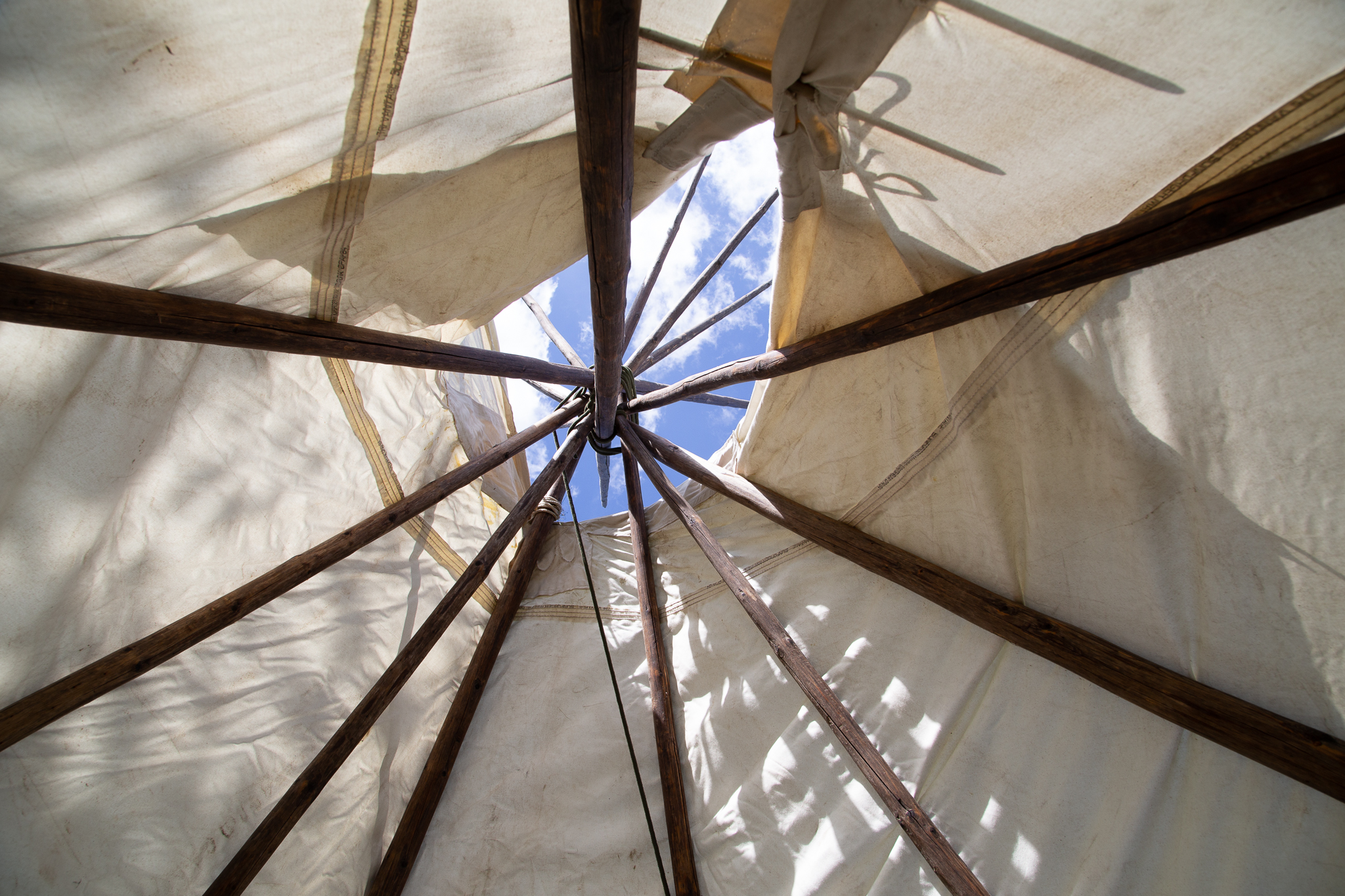 Looking up to the sky inside of a tipi. The wooden poles of the structure converge together and a gap in the white canvas shows the blue sky outside. 