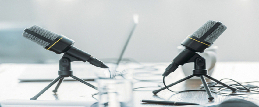 There are two wired microphones on opposite sides on the wooden table. There are glasses, a laptop, and a pen in the blurry foreground and background.