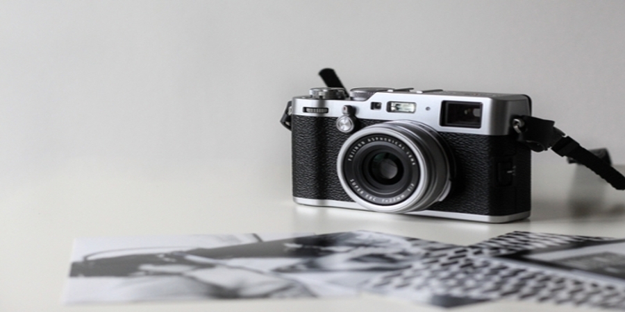 An old style camera in focus and some black and white pictures out of focus on a white table.