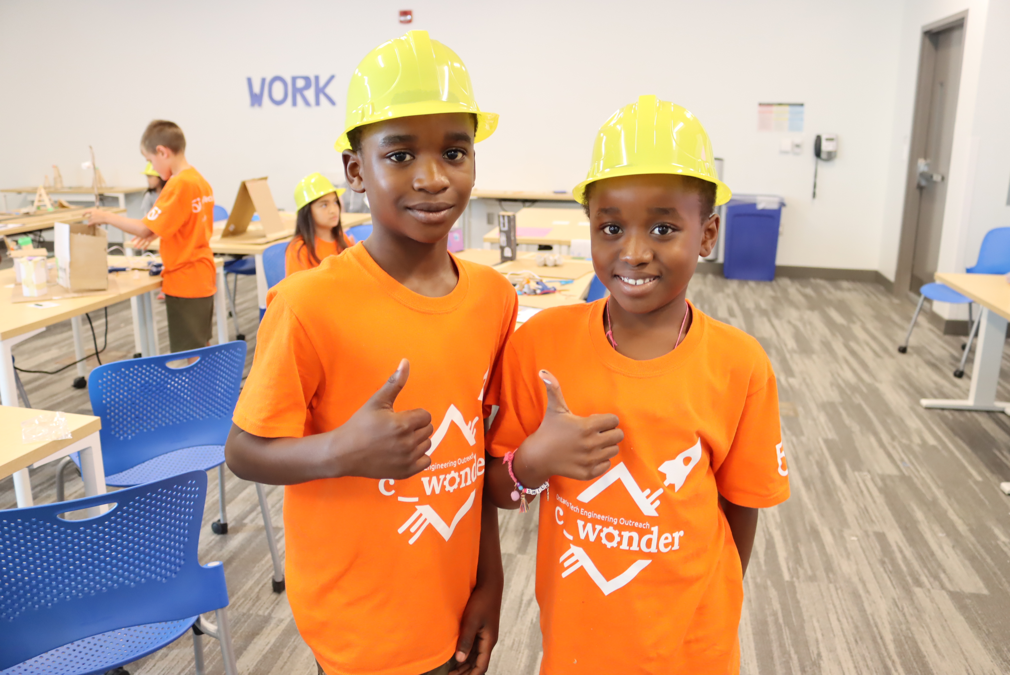 Two youth wearing construction hats and orange shirts, smiling at the camera and giving the thumbs up