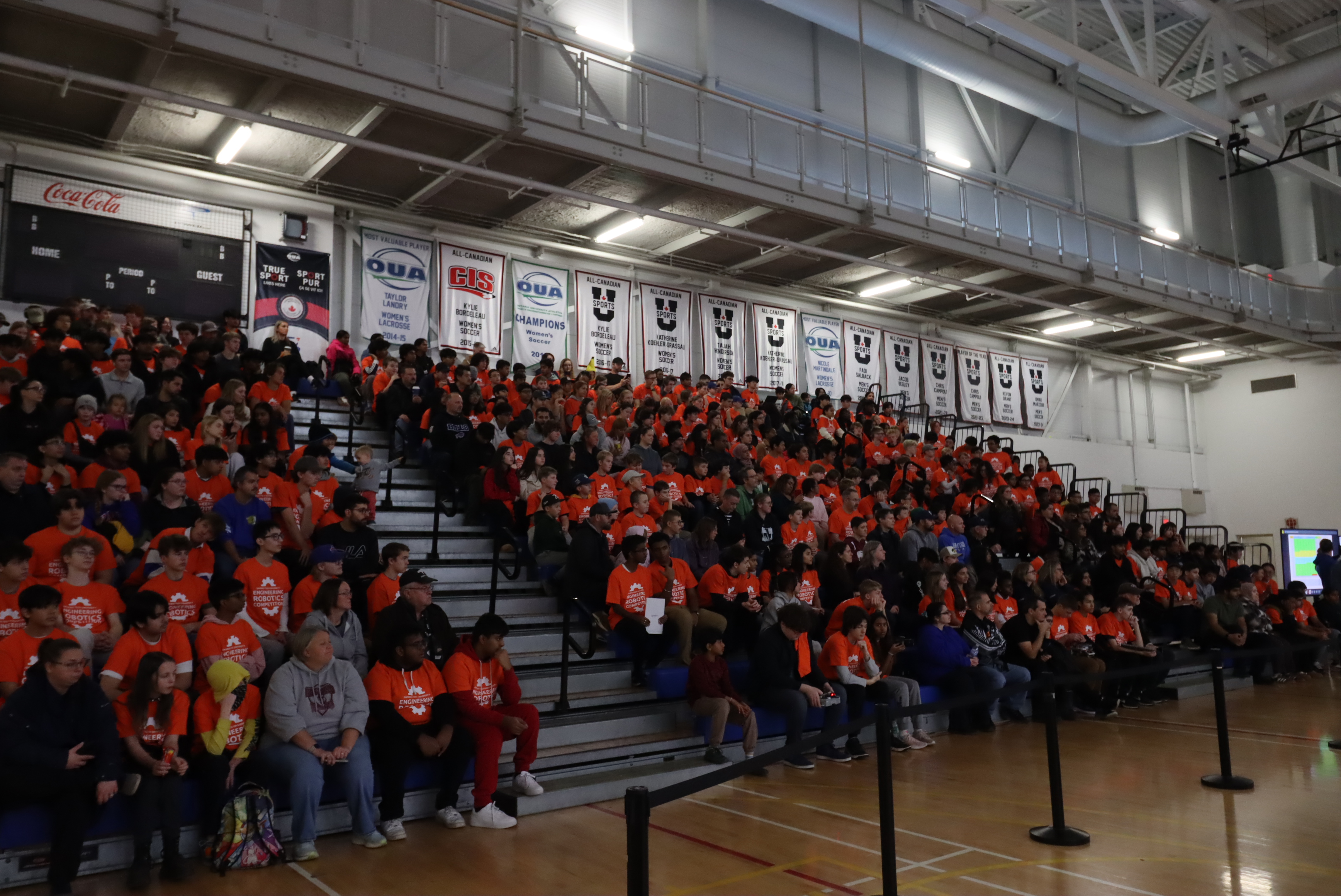 Large crowd in orange shirts seated in gym bleachers during an indoor event.