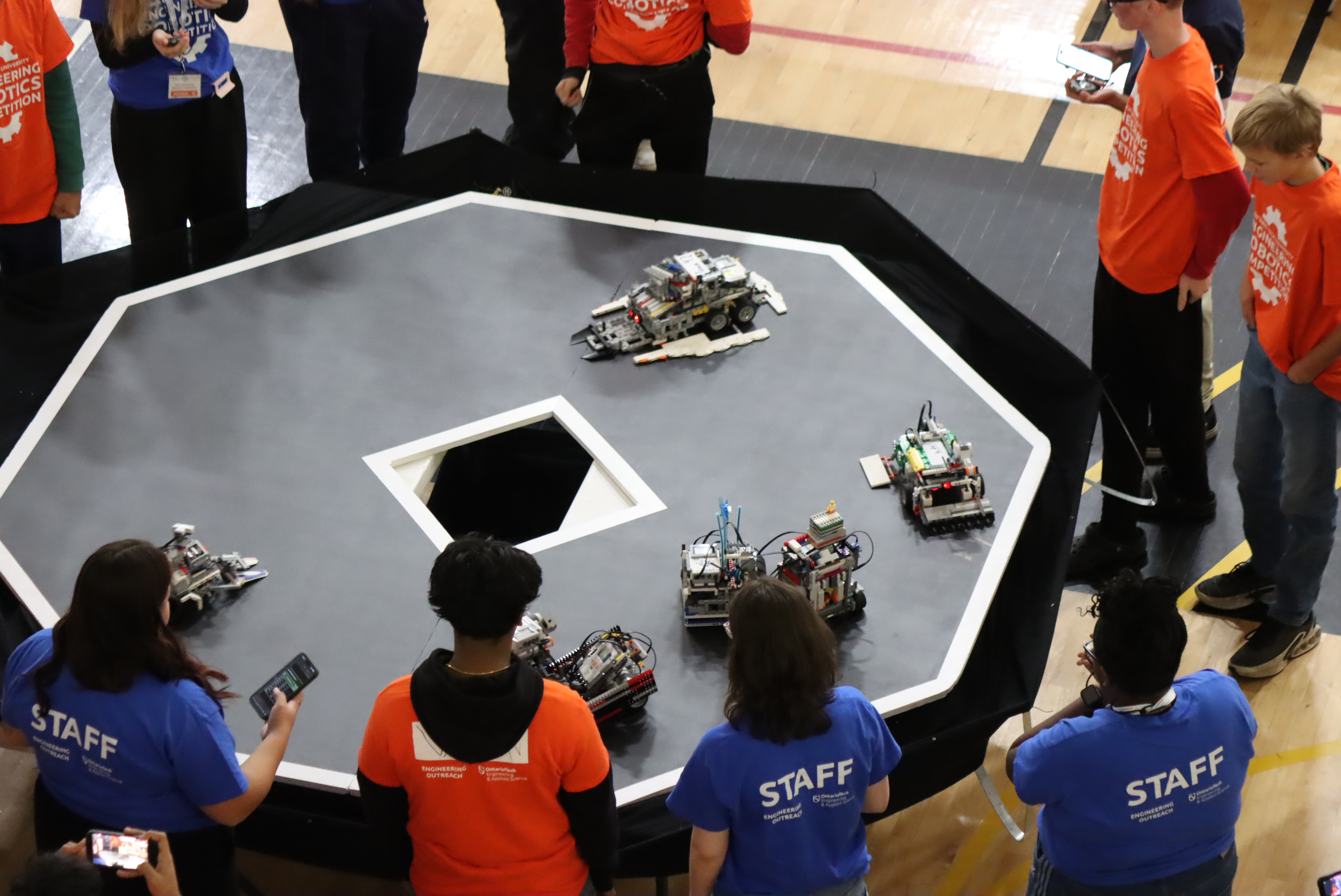 Students and staff watch several small robots compete on an octagonal arena table at a robotics competition.