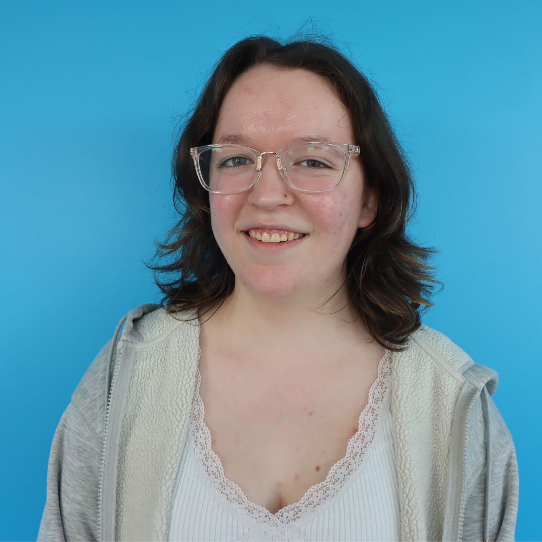 A young woman with glasses and short brown hair smiling against a bright blue background.