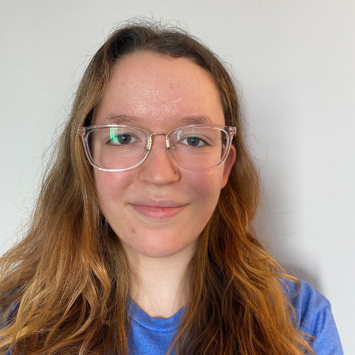 A young woman with long wavy hair wearing glasses and a blue shirt, smiling against a plain background.