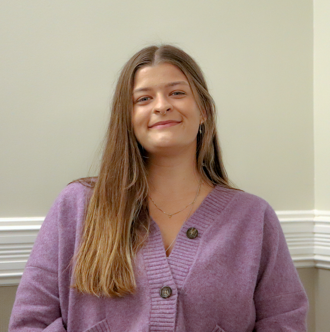 A young woman smiling and wearing a lavender cardigan, standing against a soft-colored wall.