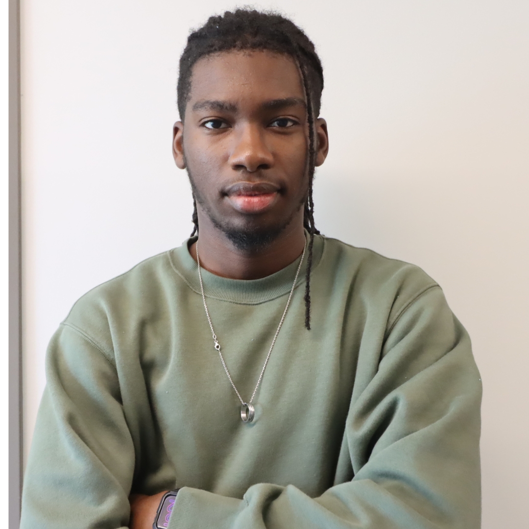 A young man with loc'd hair wearing a green sweatshirt, standing with arms crossed and a confident expression.