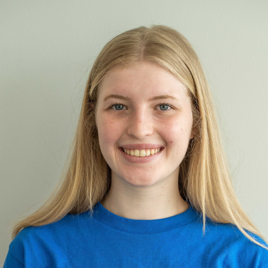 A young woman with long blonde hair smiling, wearing a blue t-shirt against a light gray background.