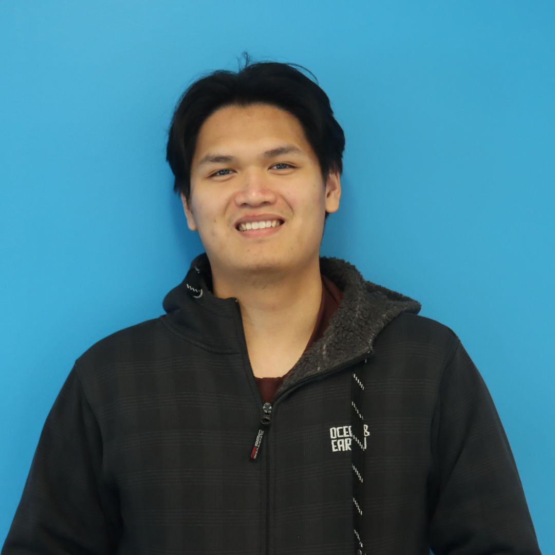 A smiling young man wearing a black jacket stands in front of a blue background.