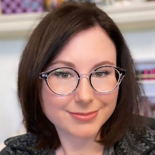 A close-up portrait of a woman with glasses, featuring a warm smile and short dark hair.