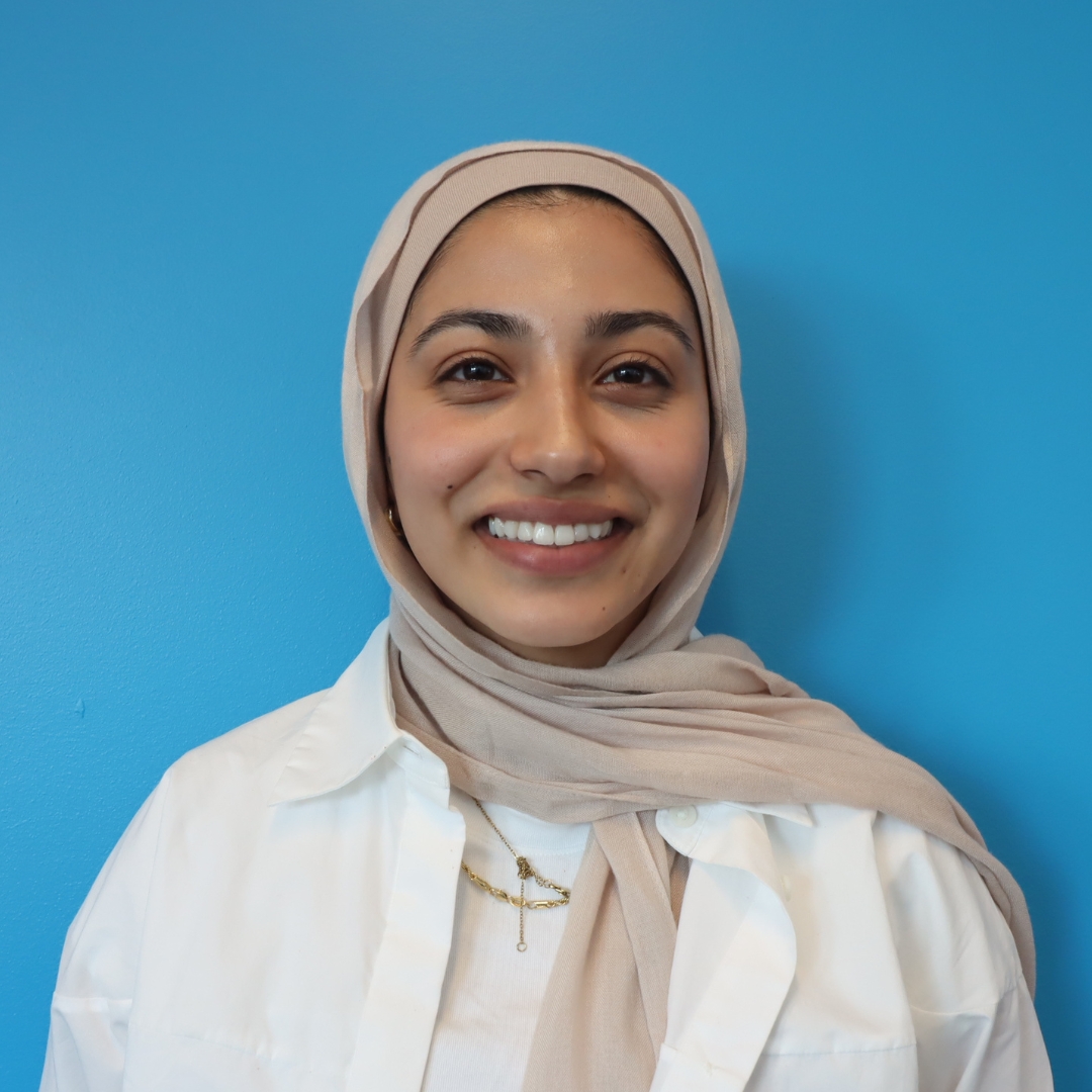 Smiling young woman wearing a hijab and a white shirt against a blue background.