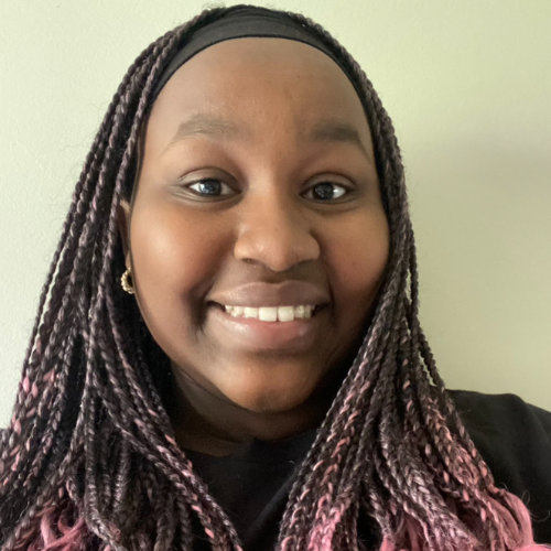 A smiling woman with braided hair featuring pink tips against a plain light background.