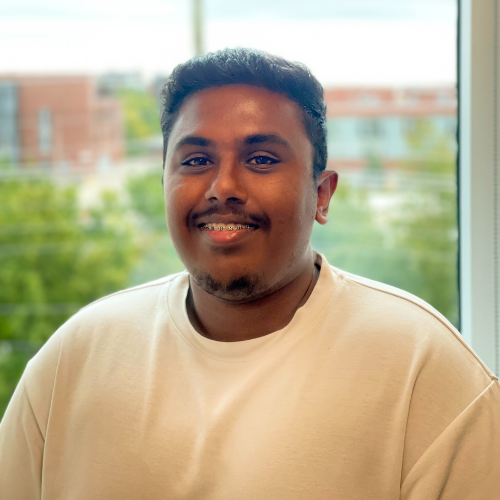 A smiling young man wearing a white sweatshirt, standing in front of a bright window with greenery in the background.