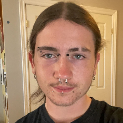 A young person with multiple facial piercings and long hair, standing in a room with light-colored walls.