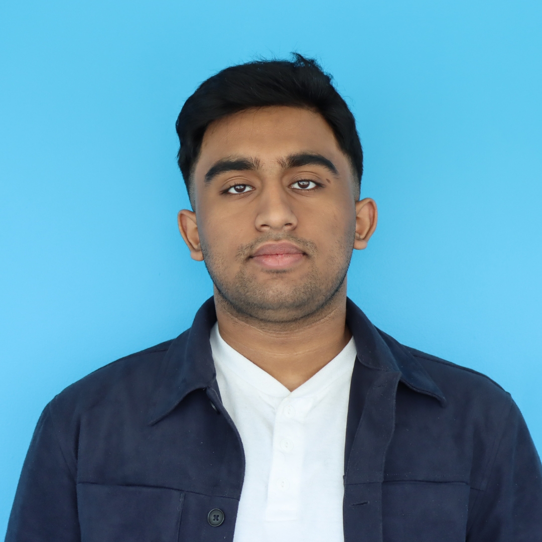 A portrait of a young man wearing a navy blue jacket over a white shirt against a bright blue background.