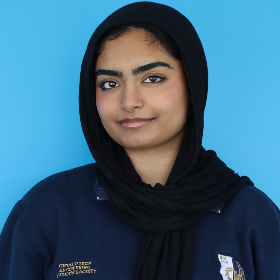 Portrait of a young woman wearing a black hijab and a blue sweater with an engineering logo against a bright blue background.
