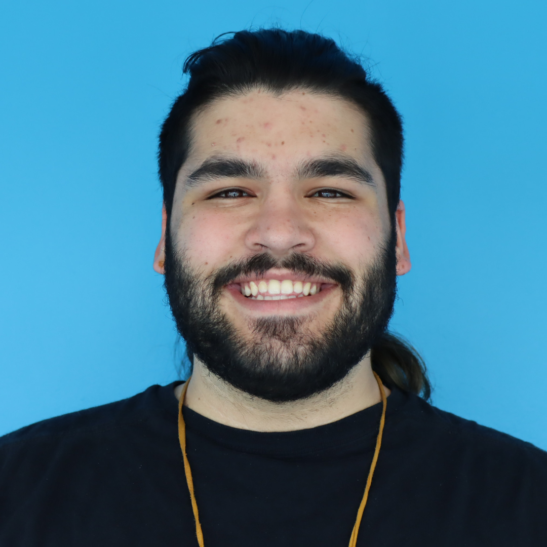 A smiling young man with a beard and long hair against a bright blue background.
