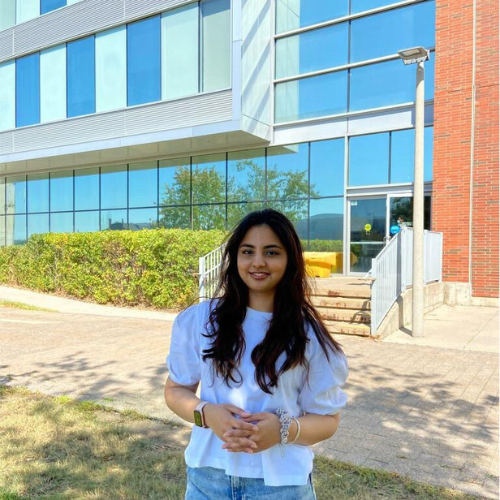 A young woman standing outdoors in front of a modern building, wearing a white blouse and jeans.