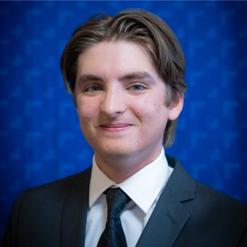 Professional headshot of a young man in a suit and tie with a blue patterned background.