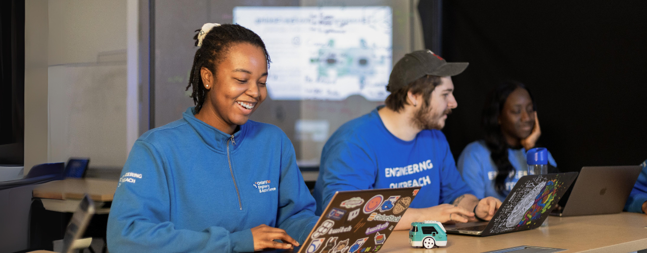 A photo of a girl in a blue sweatshirt working at a laptop in a high tech environment
