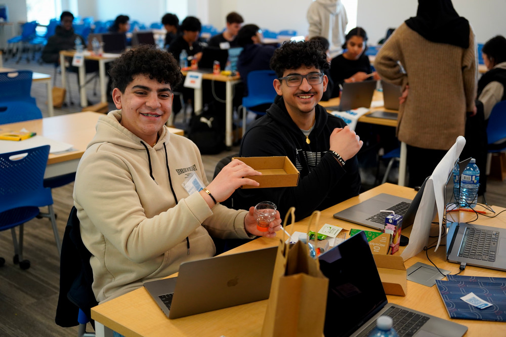 Two teens giving the thumbs up to the camera, working at laptops surrounded by technology