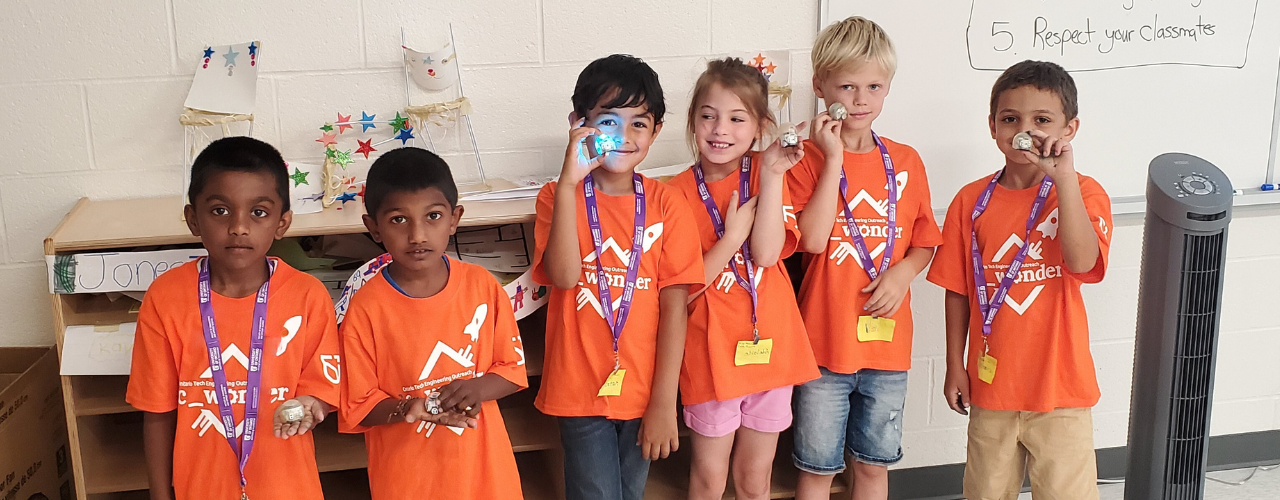 A group of children in bright orange t-shirts, smiling at the camera and golding tiny robots